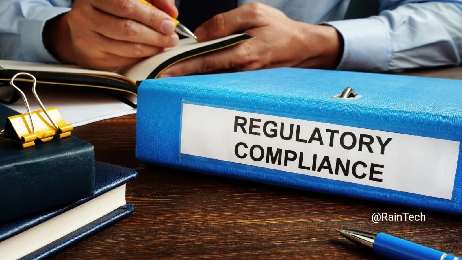 Image of a 'REGULATORY COMPLIANCE' binder and writing person, representing BPJS Indonesia costs, risks, and employer rules.