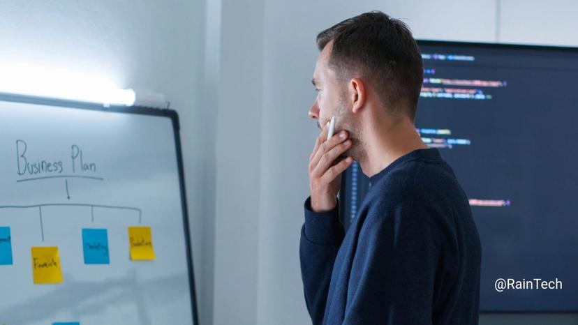 A startup founder thinking a business plan on a whiteboard with code on a screen behind him.
